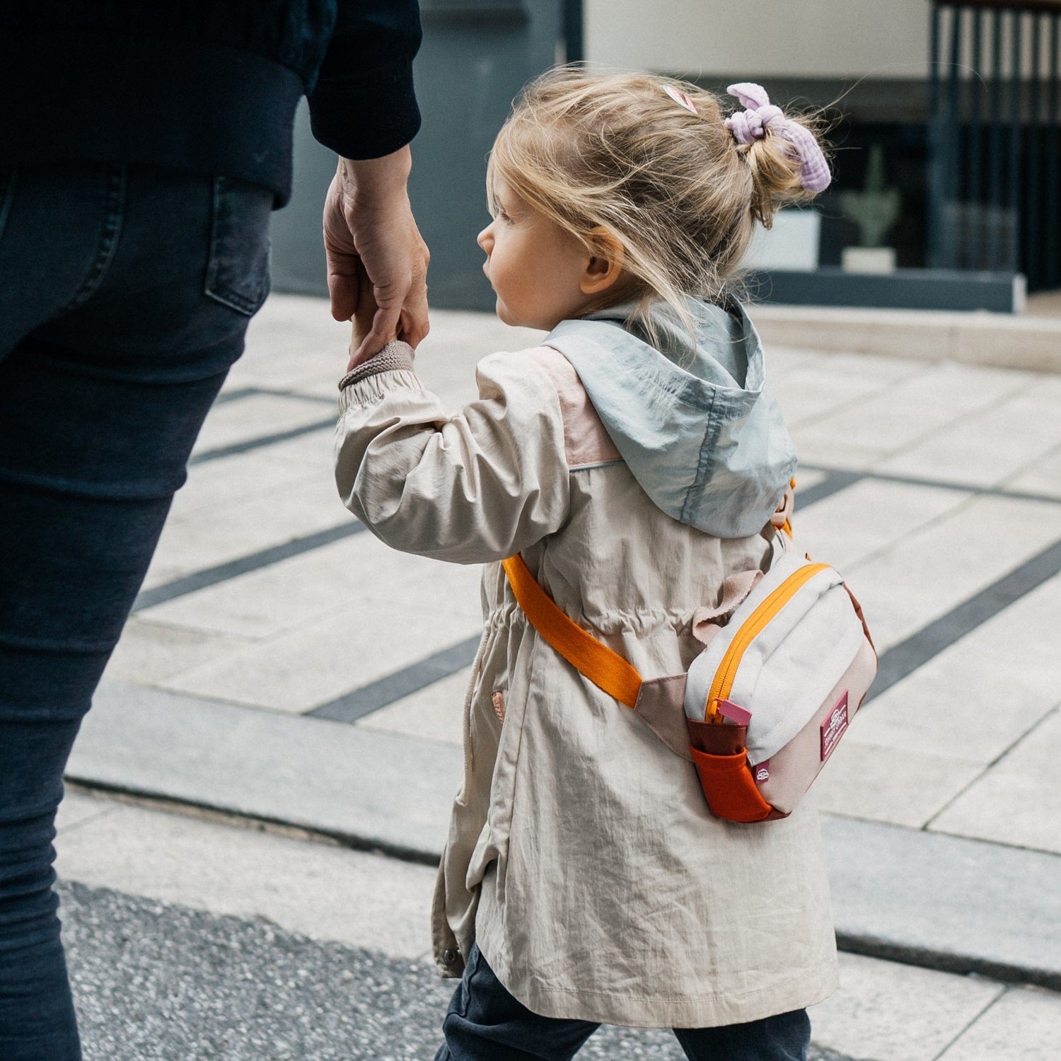 Kindermädchen mit hochgestecktem Haar hält die Hand eines Erwachsenen und trägt die Kinder Bauchtasche 'Felix' in Beige mit orangefarbenen Akzenten