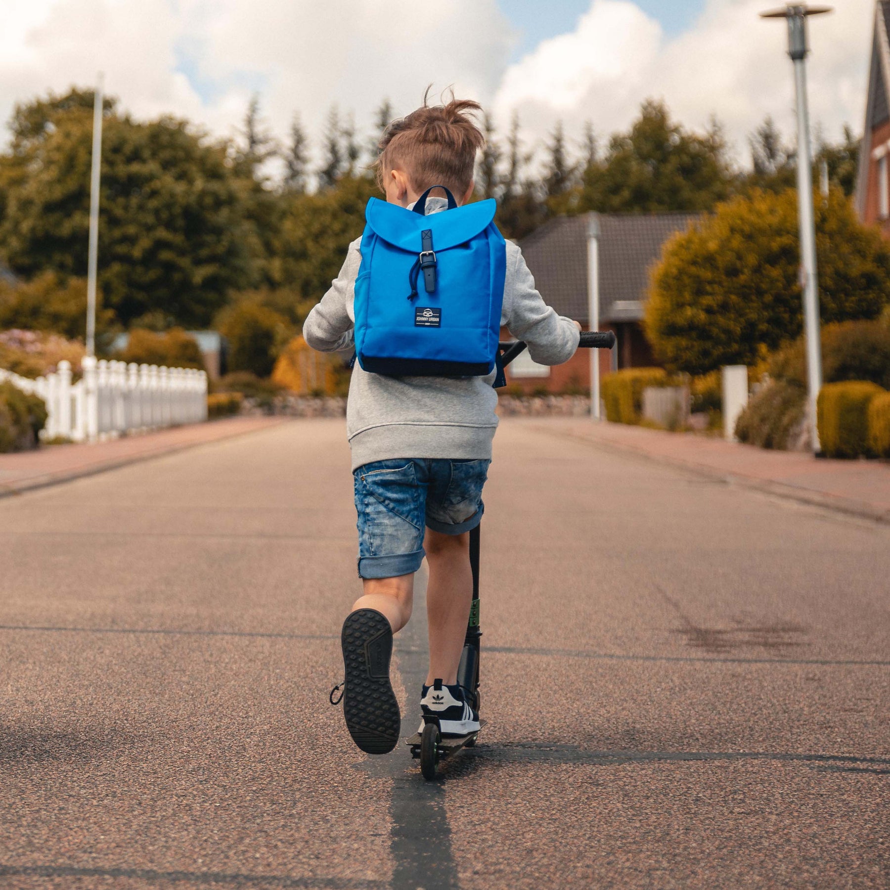 Junge mit Kinderrucksack 'Liam' in Blau fährt auf einem Roller eine Straße entlang