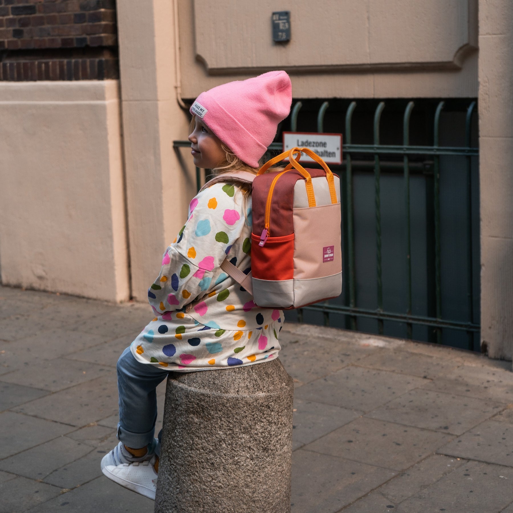 Mädchen trägt Kinderrucksack 'Linus' in Rosa mit bunten Farben, sitzt auf Mauerpfosten