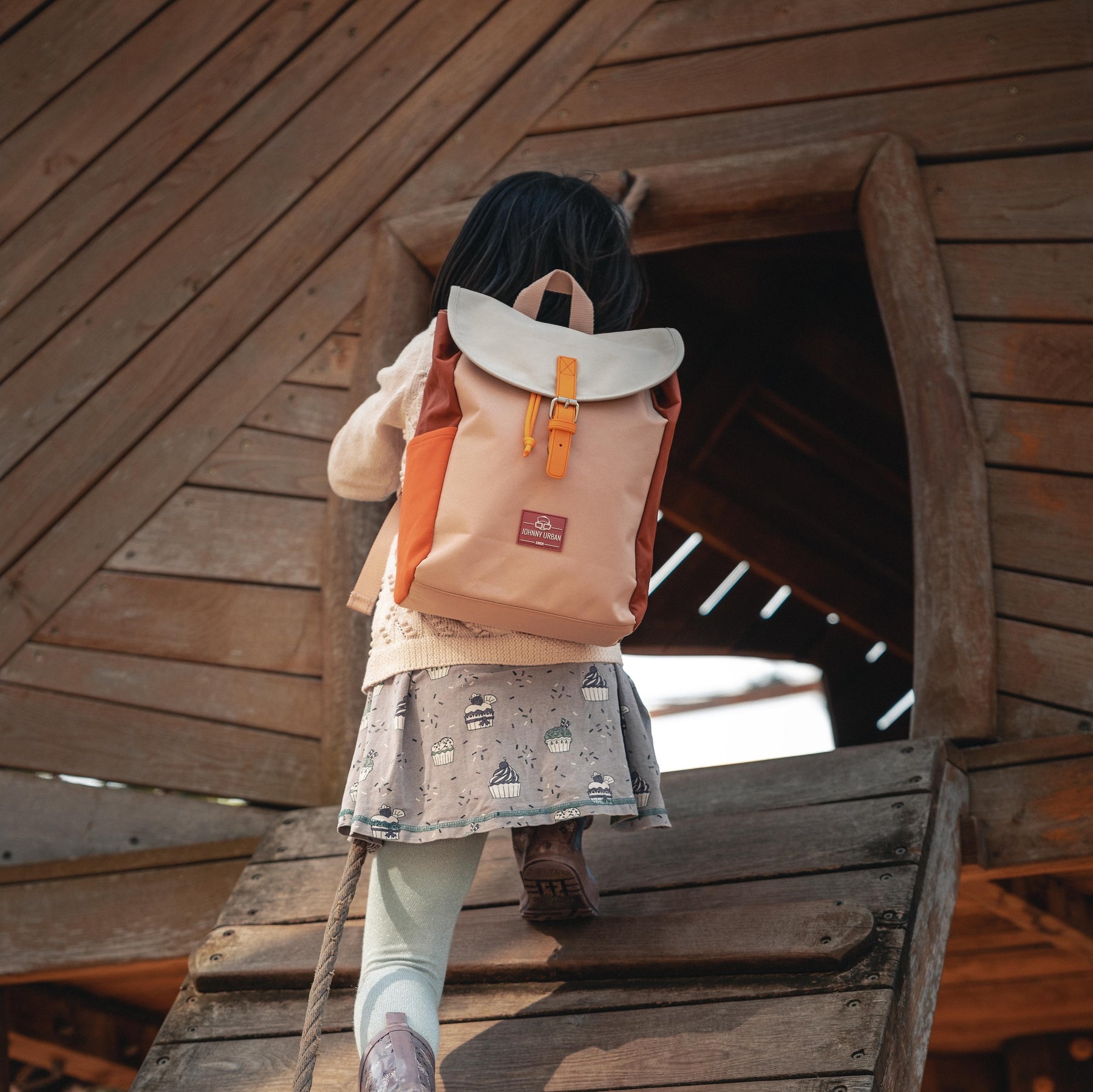 Mädchen klettert in ein Holzhaus mit rosa-buntem Kinderrucksack 'Liam' von Johnny Urban