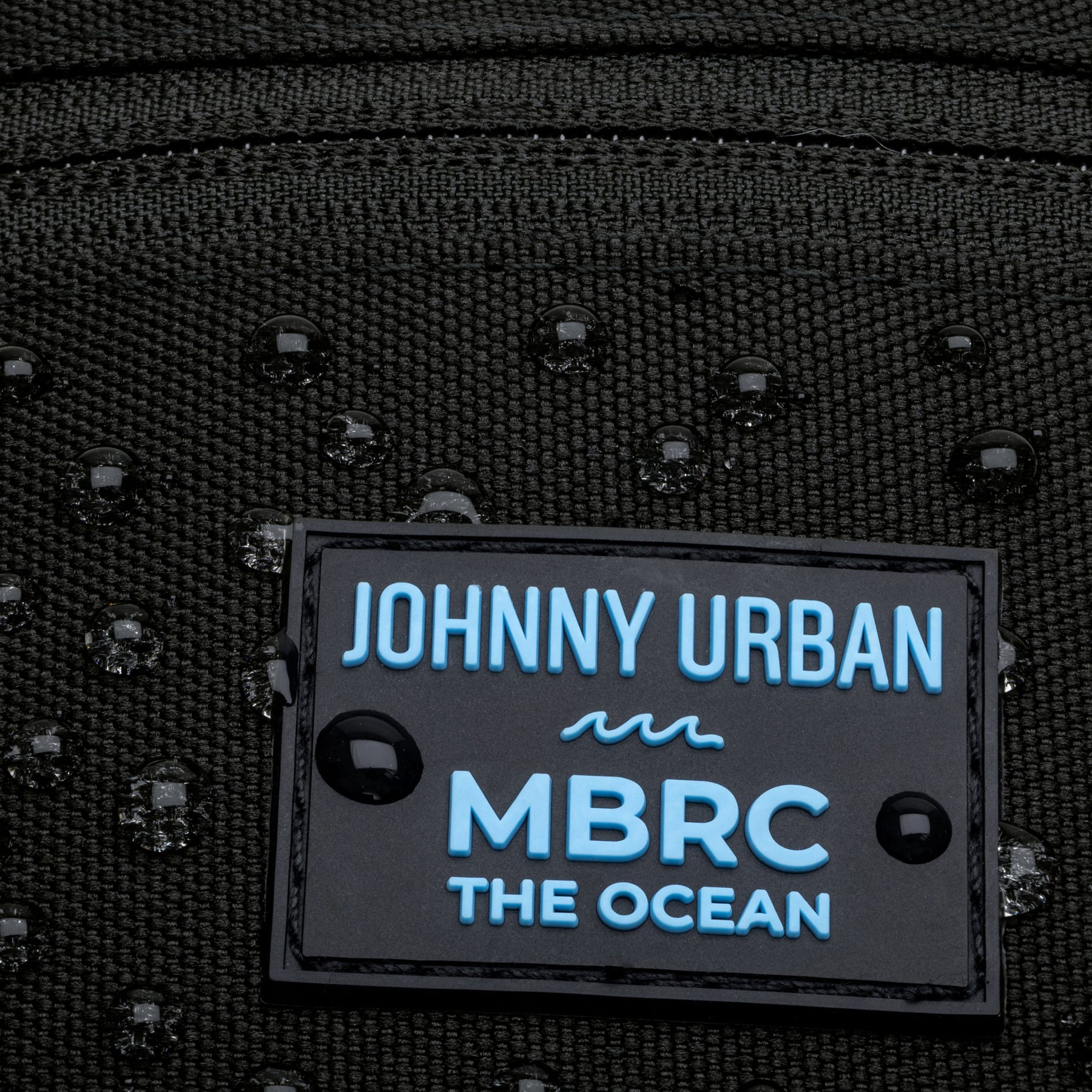 Close-up des Namensschilds 'Johnny Urban MBRC The Ocean' auf der schwarzen Bauchtasche Erik MBRC Edition mit Wassertröpfchen darauf