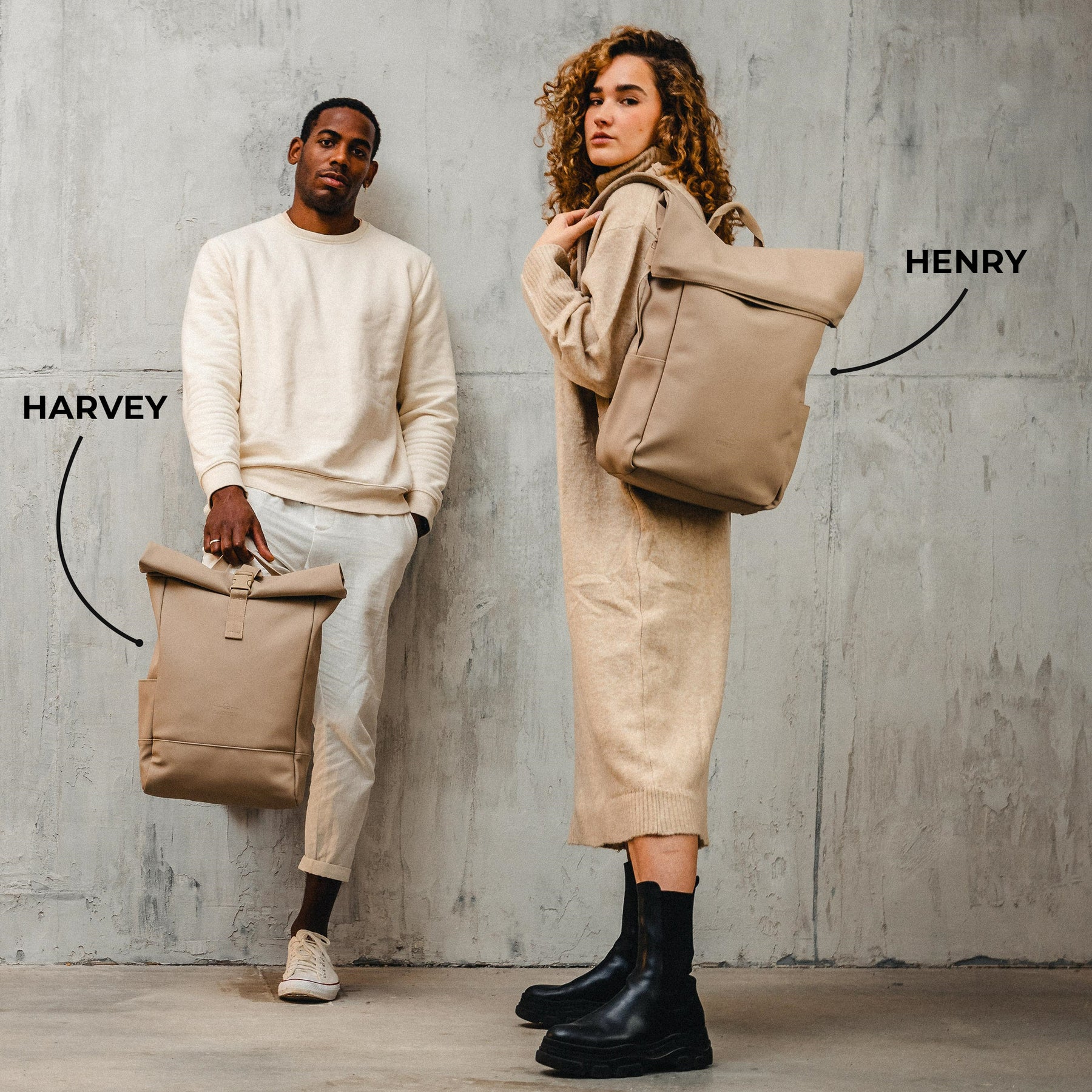 Mann hält hellbraunen Rolltop-Rucksack 'Harvey Medium', Frau trägt hellbraunen Rolltop-Rucksack 'Henry' vor grauer Betonwand