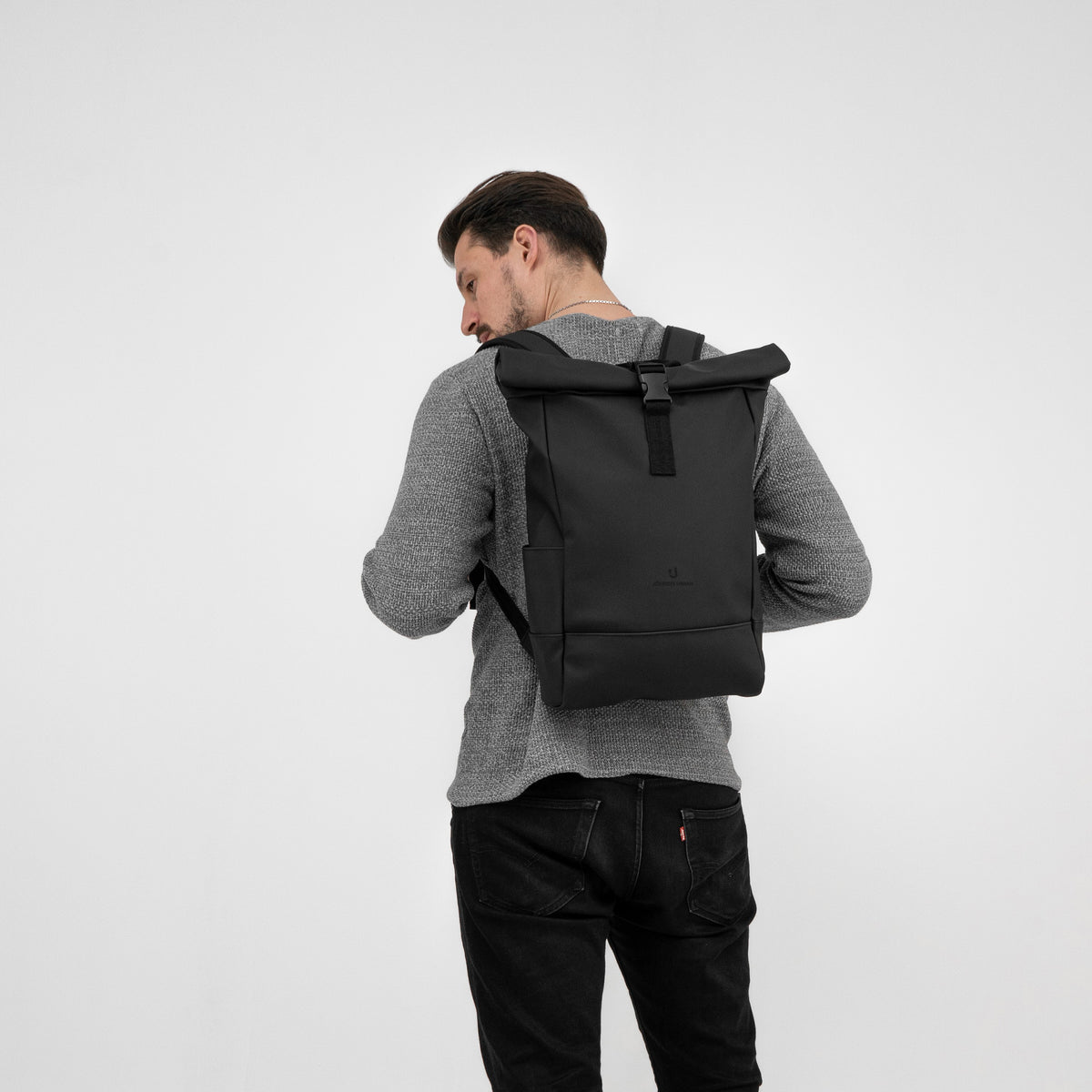 Mann von hinten trägt schwarzen Rolltop-Rucksack 'Harvey Medium' über grauem Pullover und schwarzer Jeans