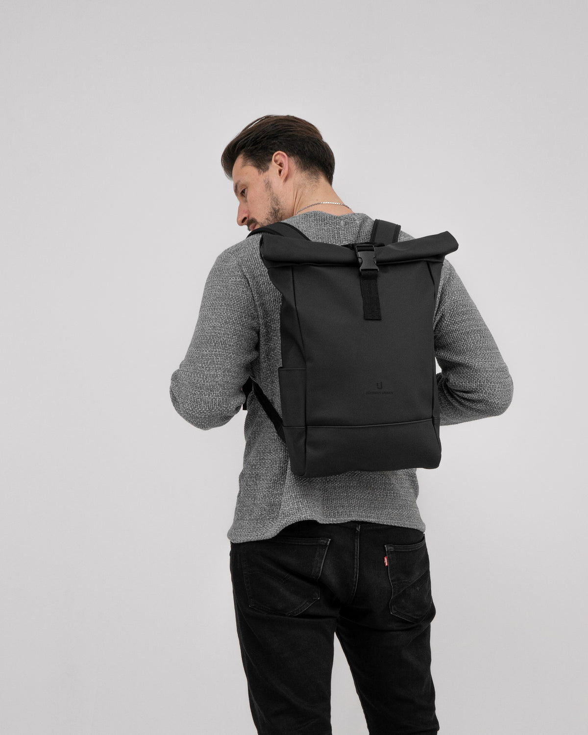 Mann von hinten trägt schwarzen Rolltop-Rucksack 'Harvey Medium' über grauem Pullover und schwarzer Jeans