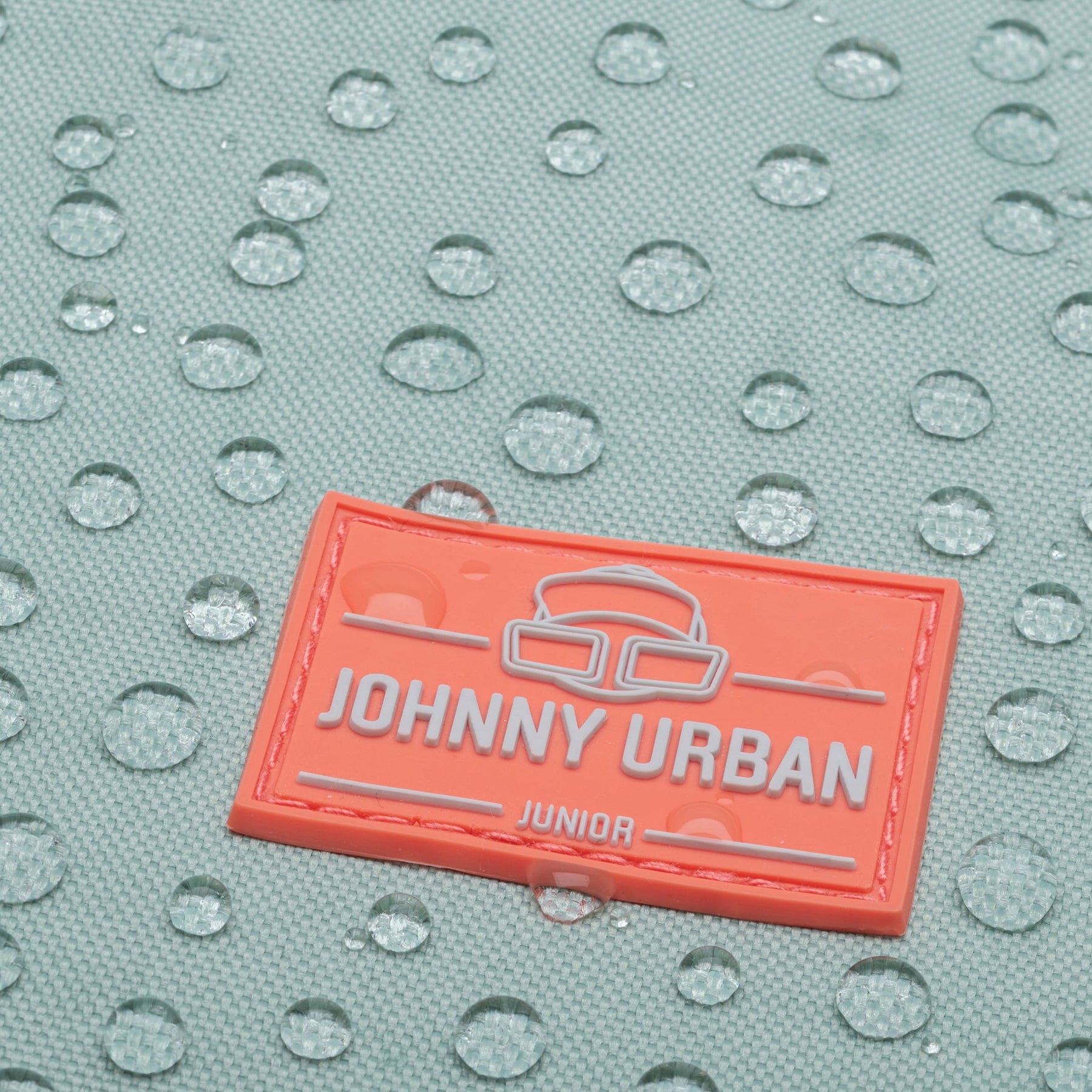 Nahaufnahme des wasserabweisenden Materials des hellblauen Rolltop Kinderrucksacks 'Aaron' mit einem orangefarbenen Johnny Urban Junior Logo.