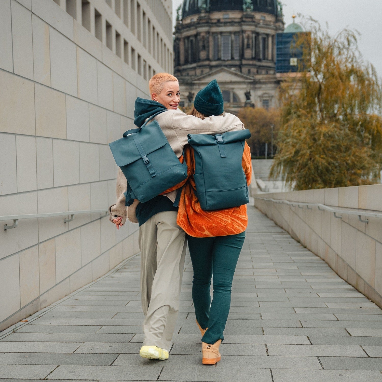 Zwei Frauen laufen Arm in Arm in der Stadt, tragen petrolfarbene Rolltop-Rucksäcke 'Harvey Medium' von Johnny Urban