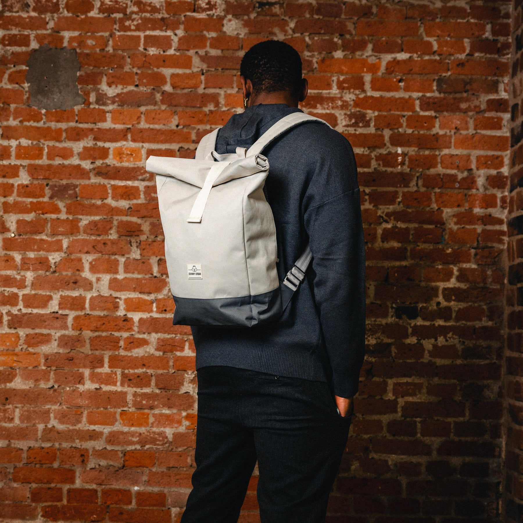 Mann trägt Rolltop-Rucksack 'Ryan' in Beige mit dunkelgrauem Boden vor Backsteinwand