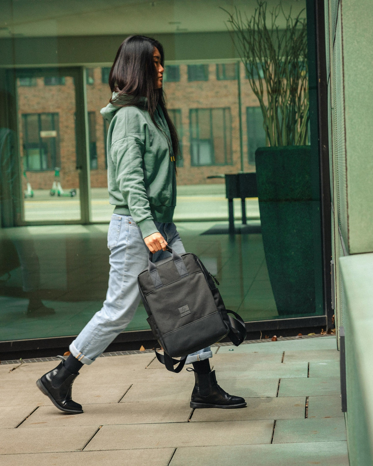 Frau trägt schwarzen Rucksack 'Jona Large' von Johnny Urban in der Hand, geht an Glasfassade vorbei