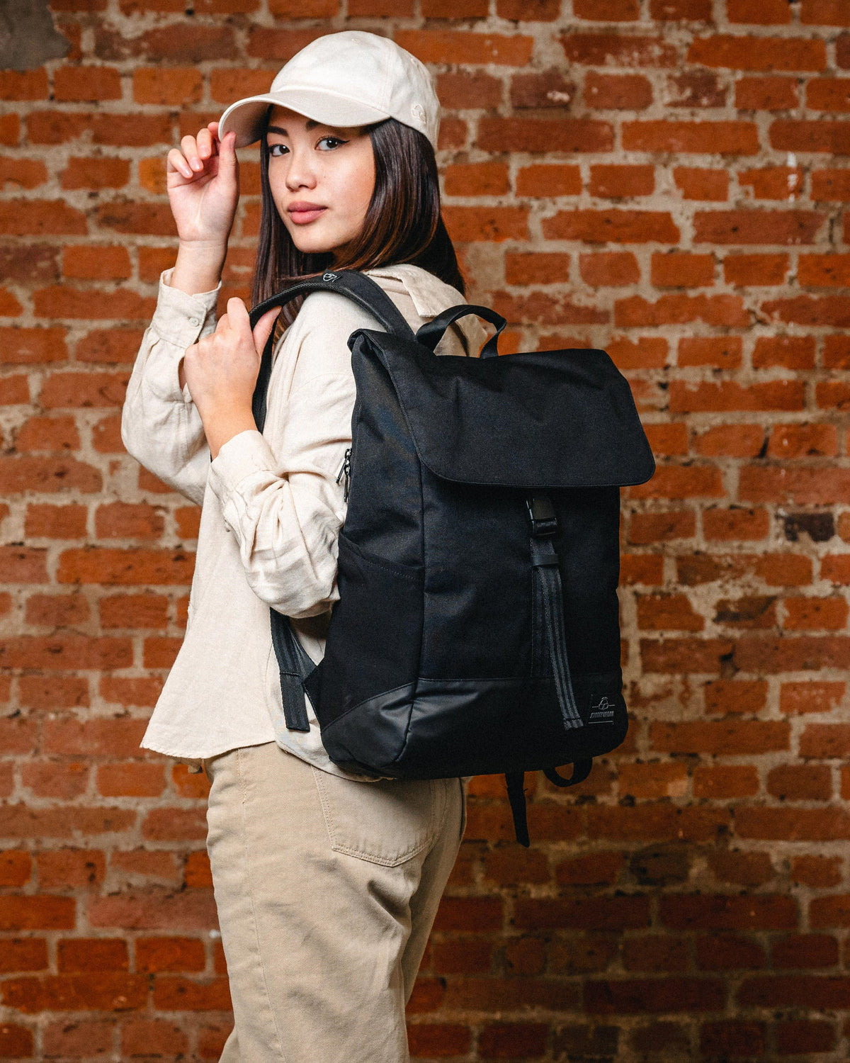 Frau mit schwarzem Rucksack 'Mika', hellbeige Cap und beiger Kleidung vor roter Ziegelwand