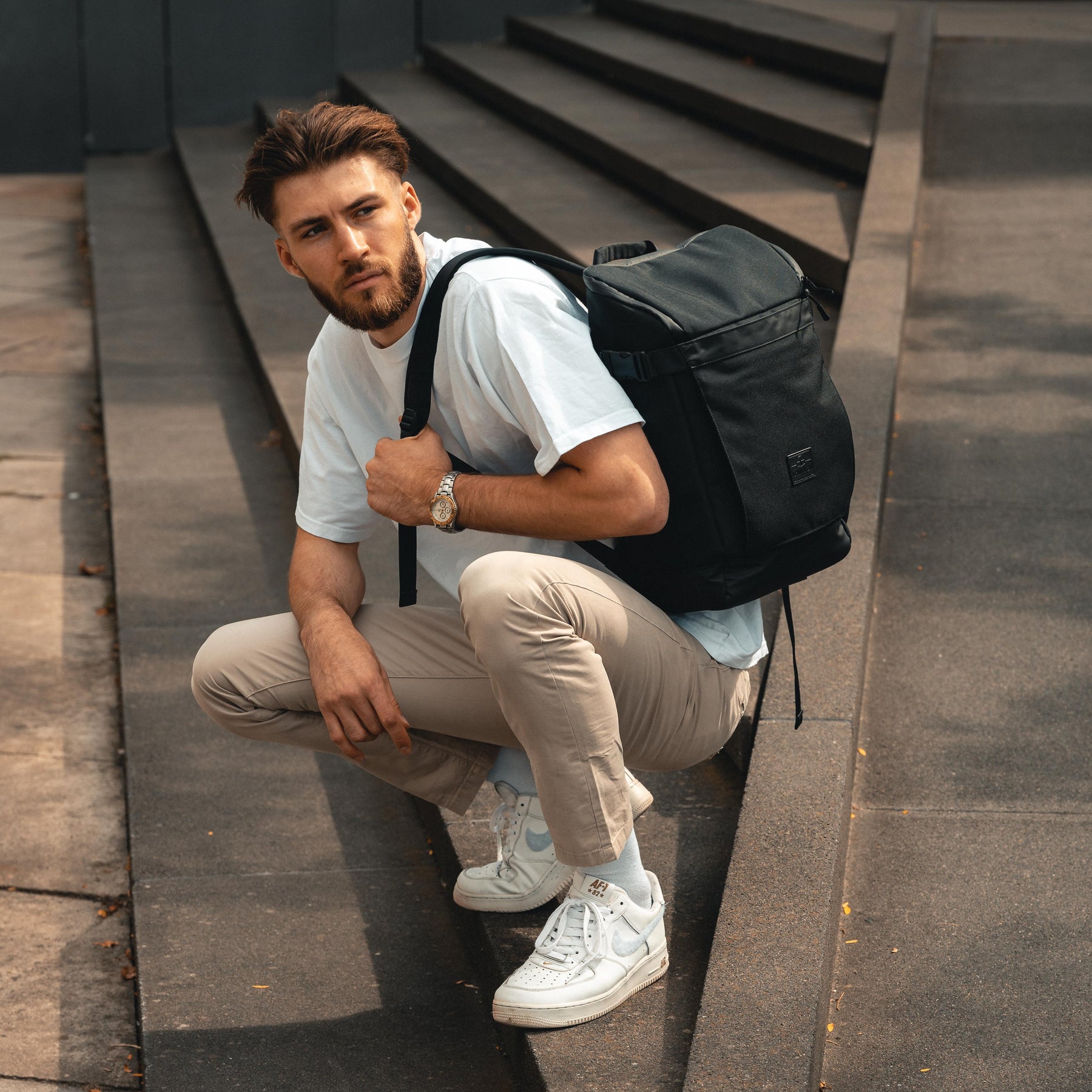 Mann mit weißem T-Shirt, beige Hose und schwarzem Rucksack 'Luca' sitzt auf einer Außentreppe