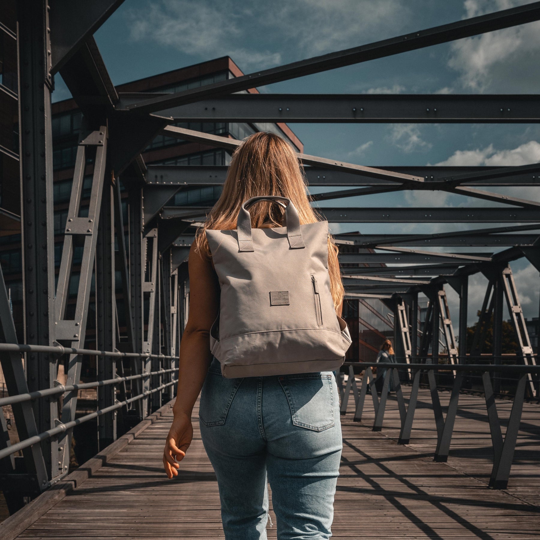 Frau trägt beige Rucksacktasche 'Ivy' von Johnny Urban auf Holzbrücke im urbanen Umfeld