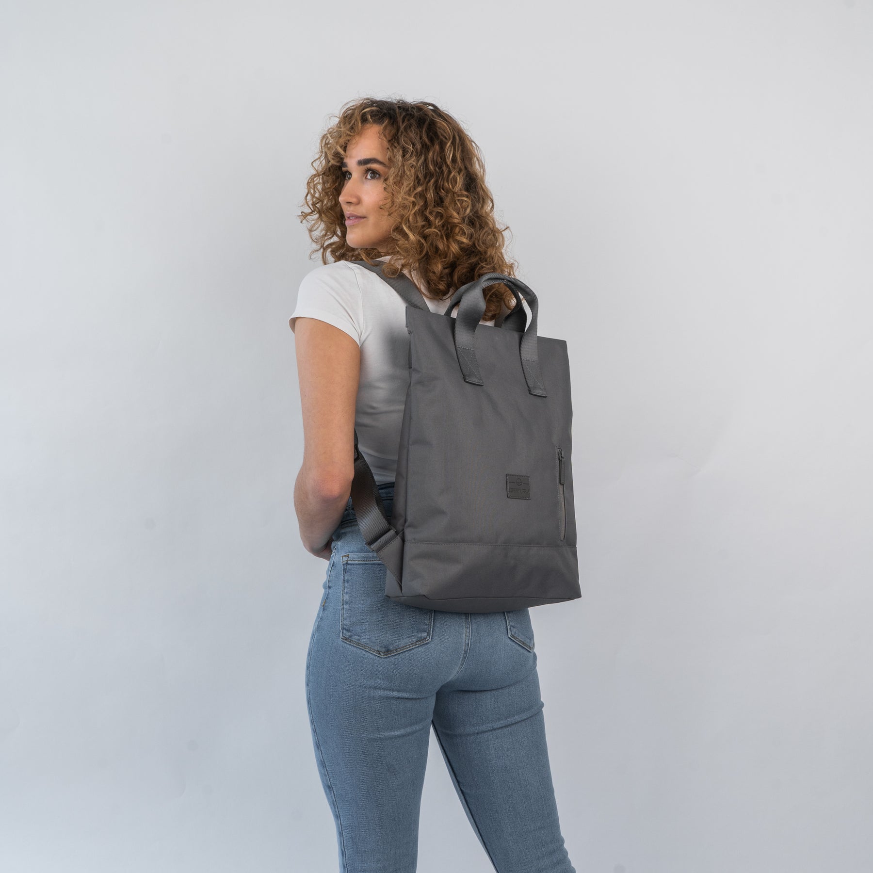 Frau mit lockigem Haar trägt dunkelgrauen Rucksacktasche 'Ivy' von Johnny Urban über weißen T-Shirt und Jeans