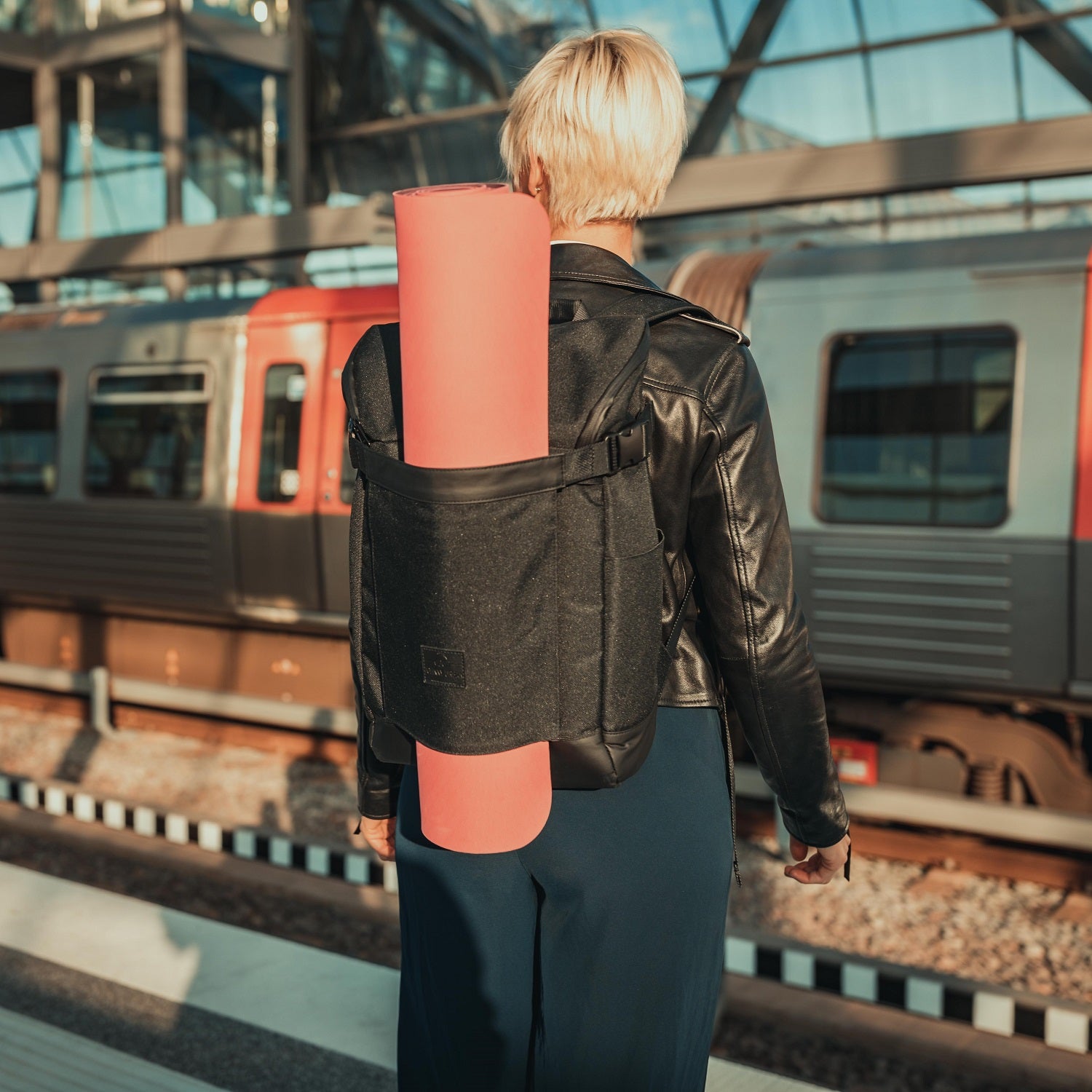 Frau mit blondem Kurzhaar trägt schwarzen Rucksack "Luca" mit rosafarbener Yogamatte am Bahnhof