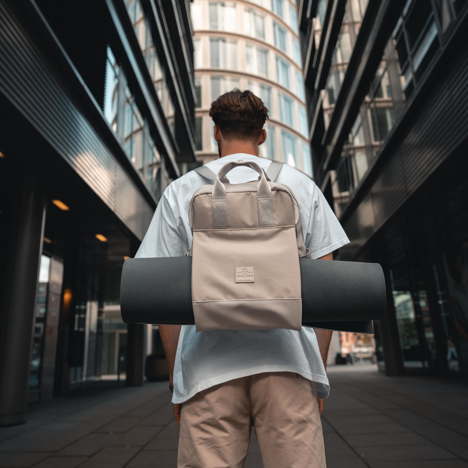 Mann mit beigem Rucksack 'Jona Large' trägt schwarze Yogamatte in urbaner Umgebung