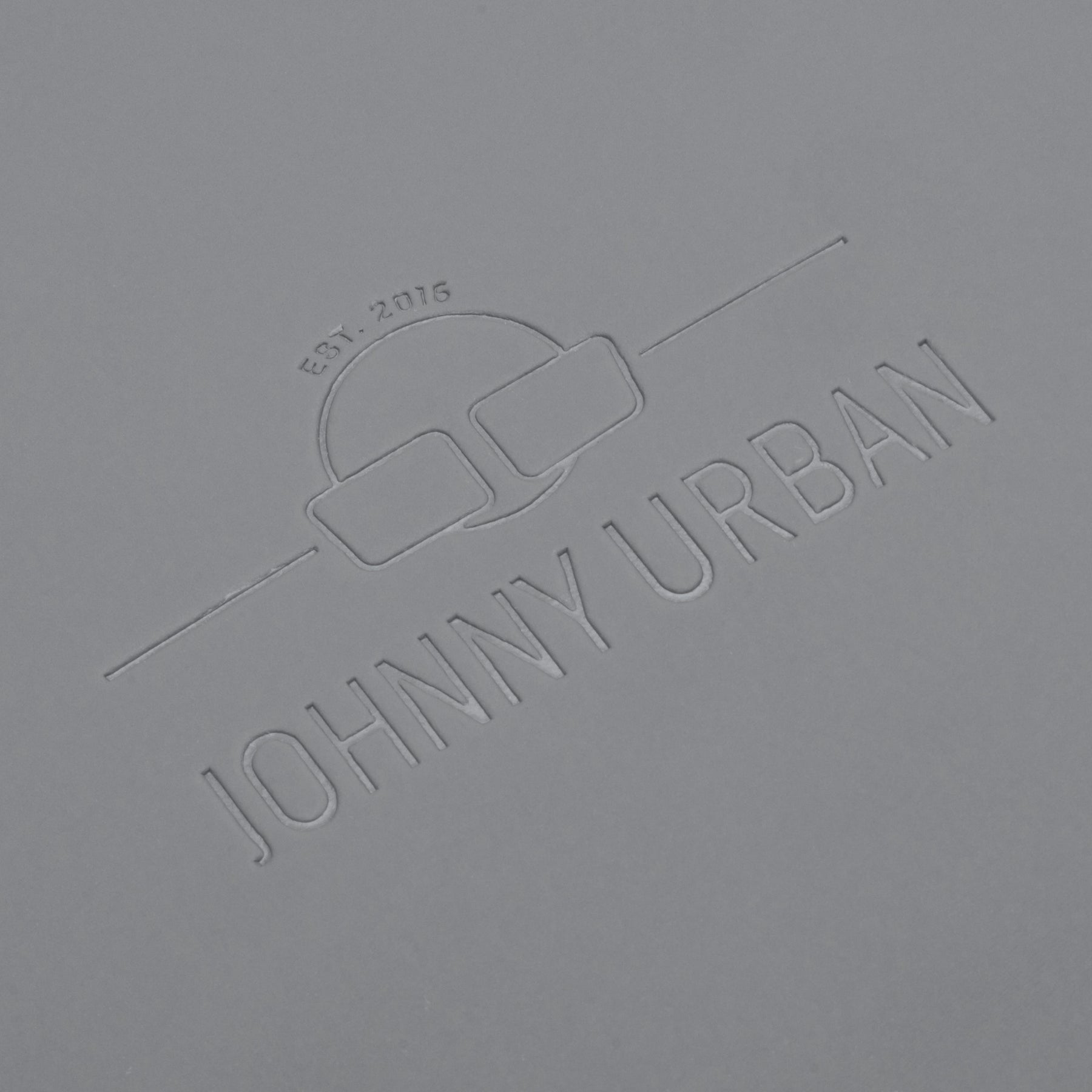 Geprägtes Johnny Urban Logo mit Sonnenbrillensymbol und Text 'Est. 2016' auf grauem Material