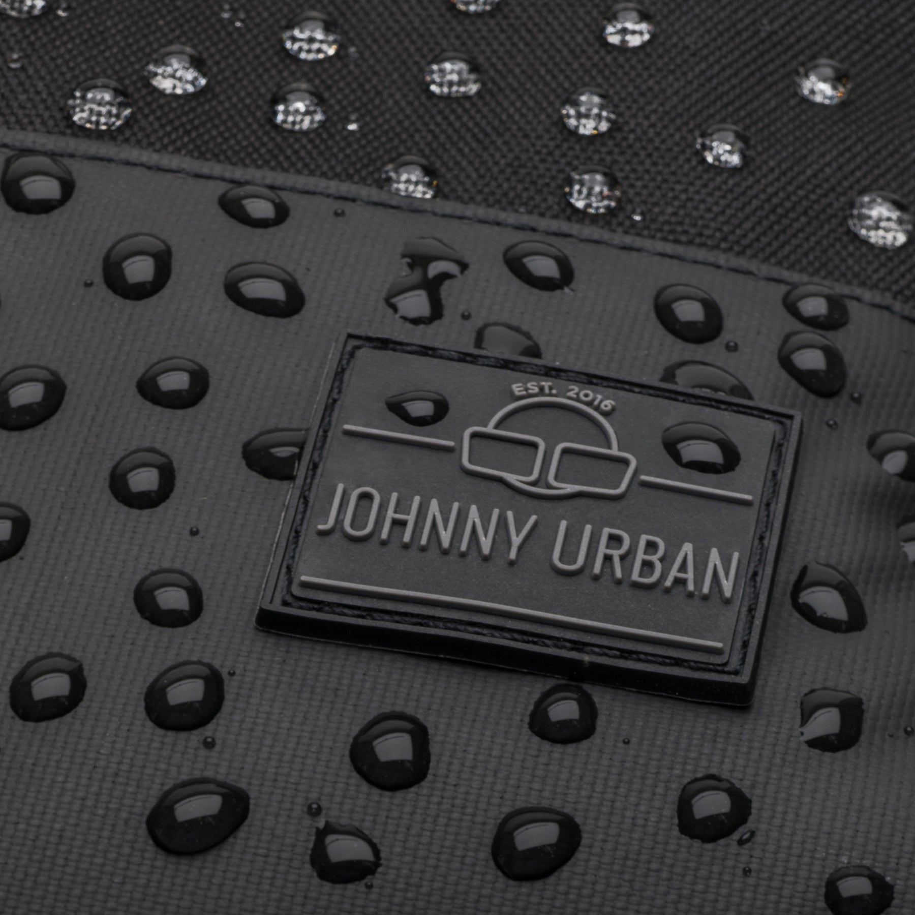 Nahaufnahme des wasserdichten schwarzen Rucksacks Mika von Johnny Urban mit Wassertropfen auf der Oberfläche und Markenlogo