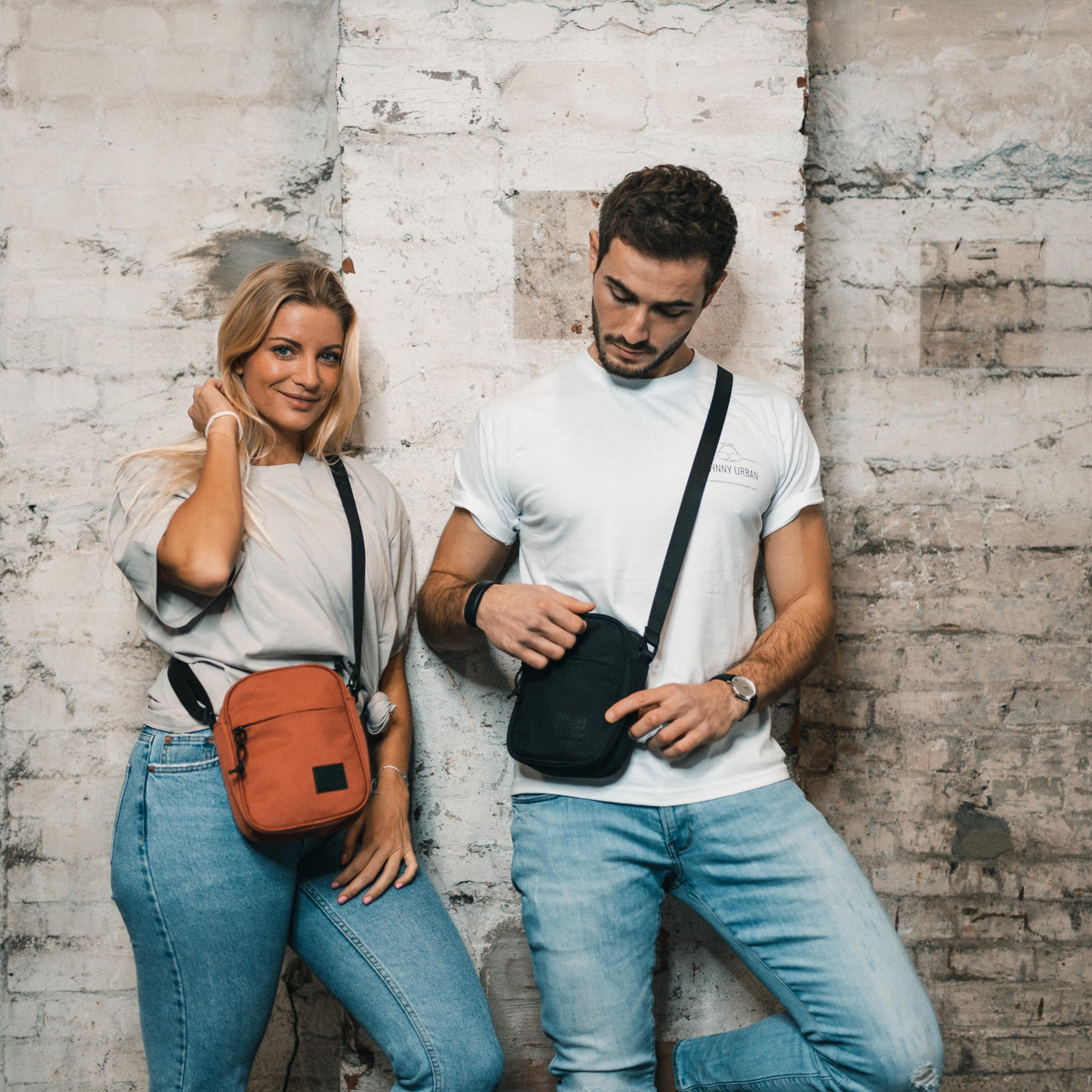 Frau und Mann lehnen an Wand, tragen Umhängetasche 'Luis' in Orange und Schwarz, lässige Kleidung