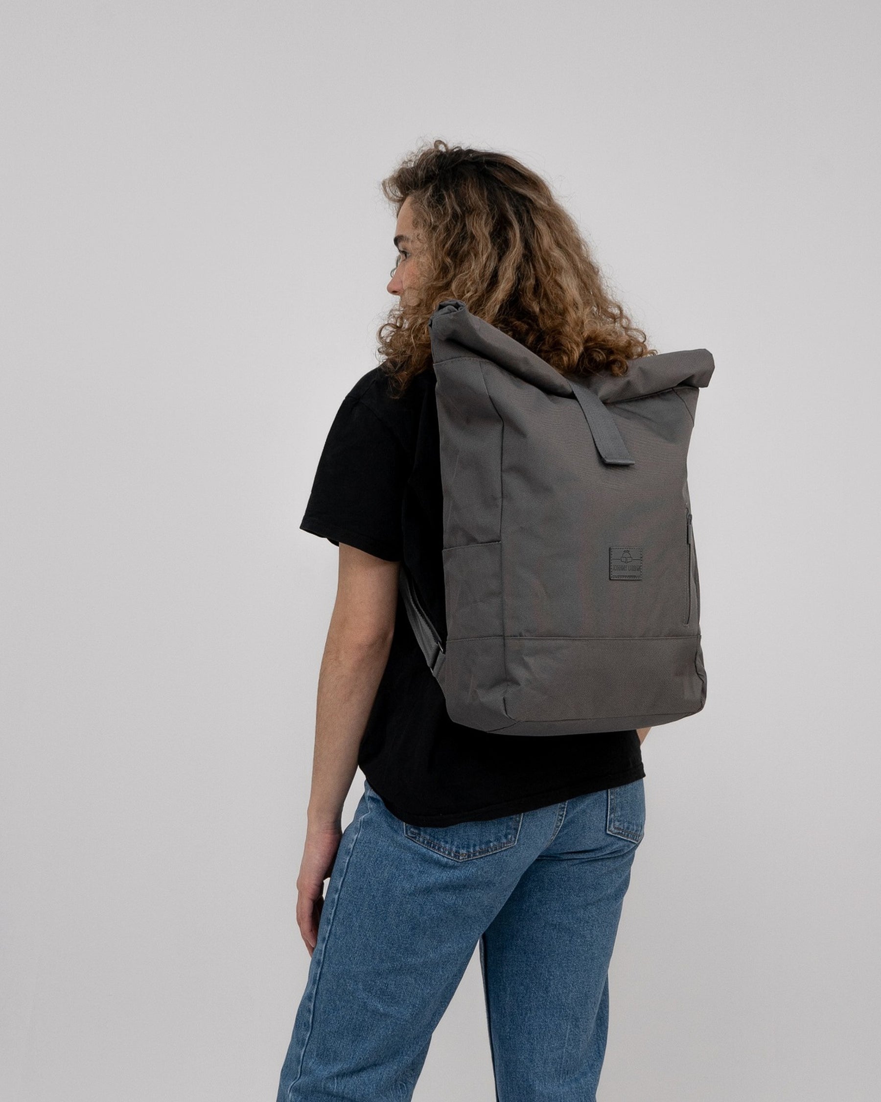 Dunkelgrauer Rolltop-Rucksack 'Robin Large' von Johnny Urban an einer Frau, seitlich stehend, mit lockigen Haaren, schwarzem T-Shirt und blauen Jeans