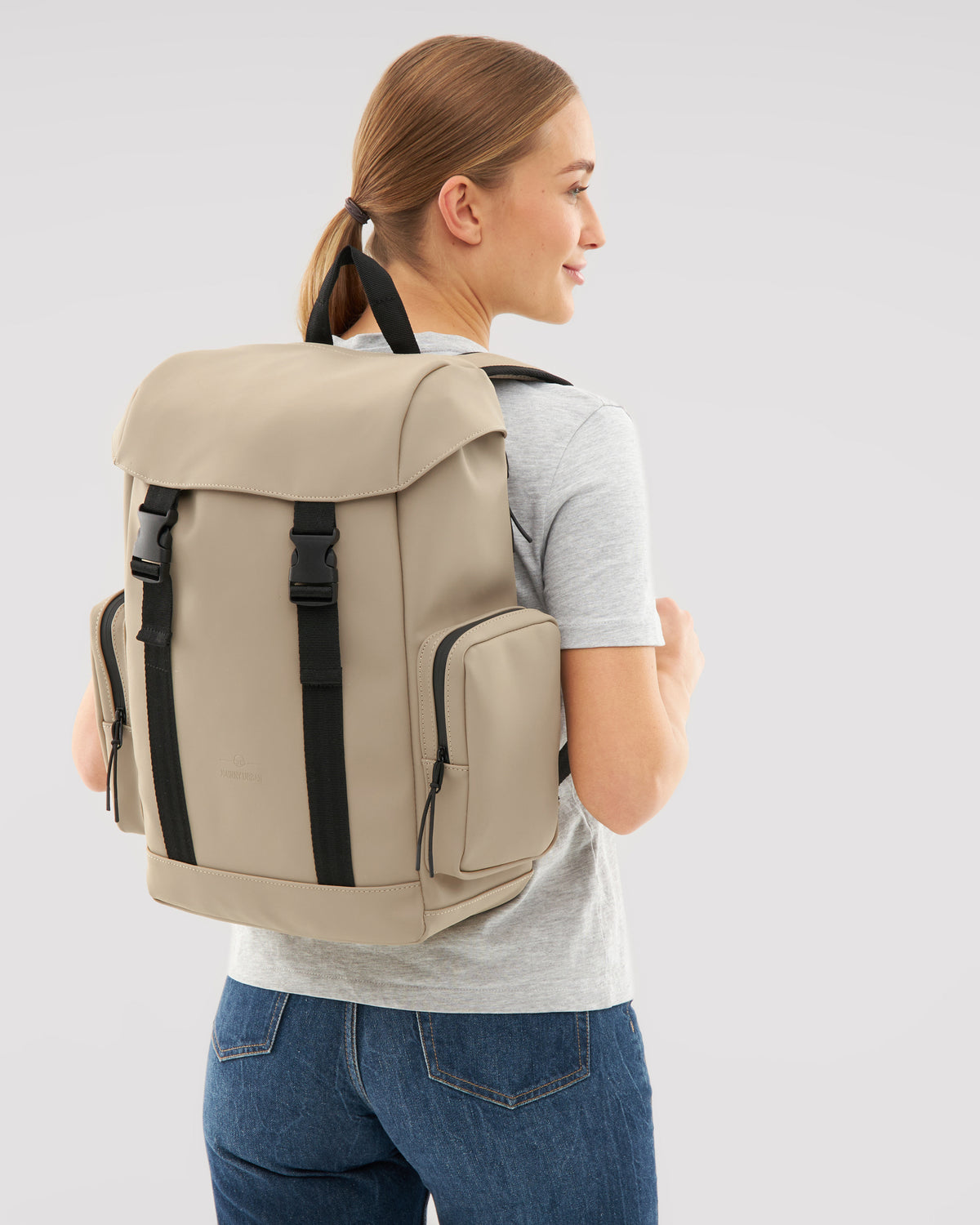 Beige Rucksack 'Finley' mit schwarzen Verschlüssen, getragen von Frau in grauem T-Shirt und blauer Jeans